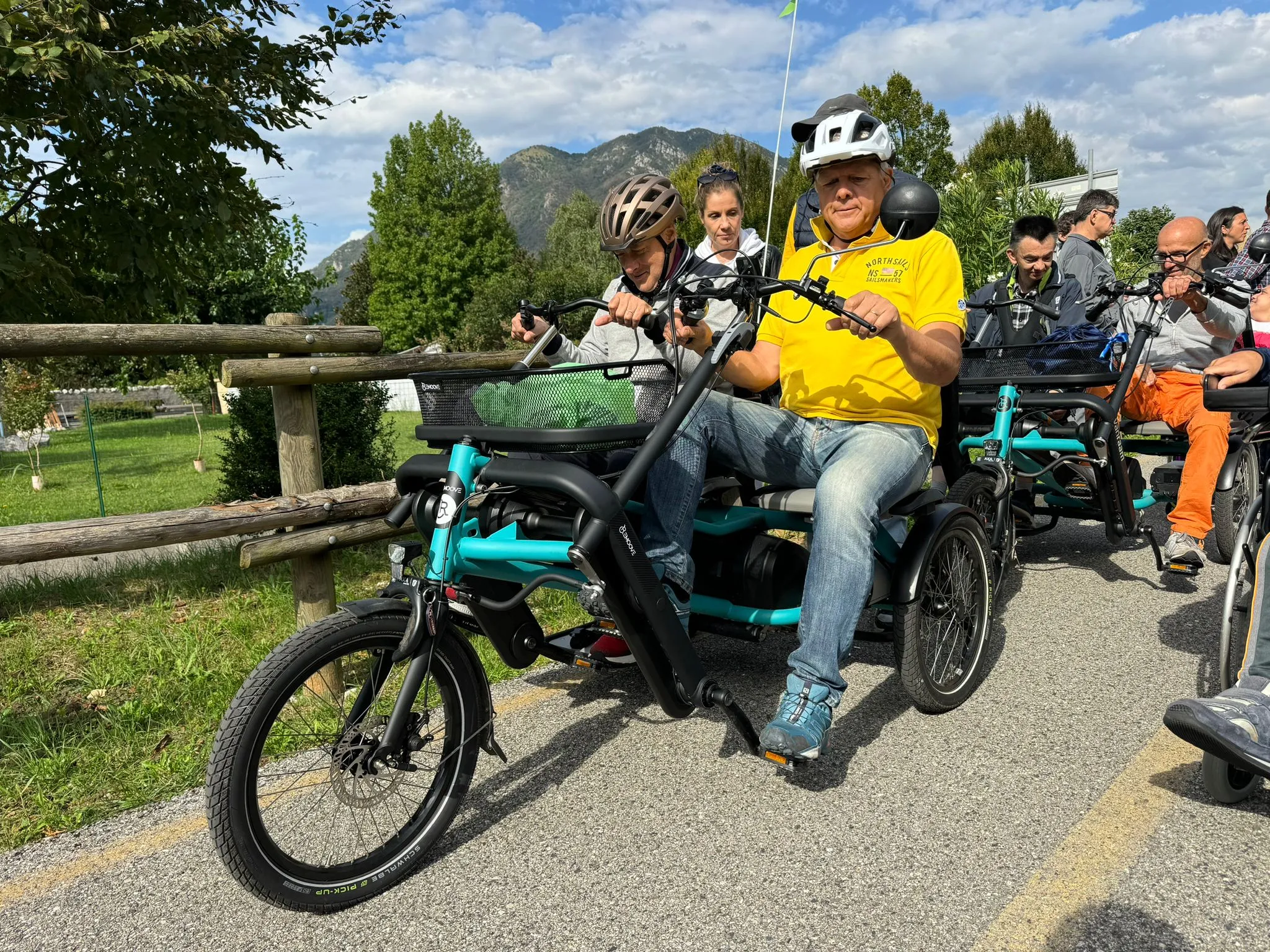 Un gruppo di ciclisti e persone su special bike si ritrova all'aperto prima di un'uscita, con le montagne del Friuli sullo sfondo.