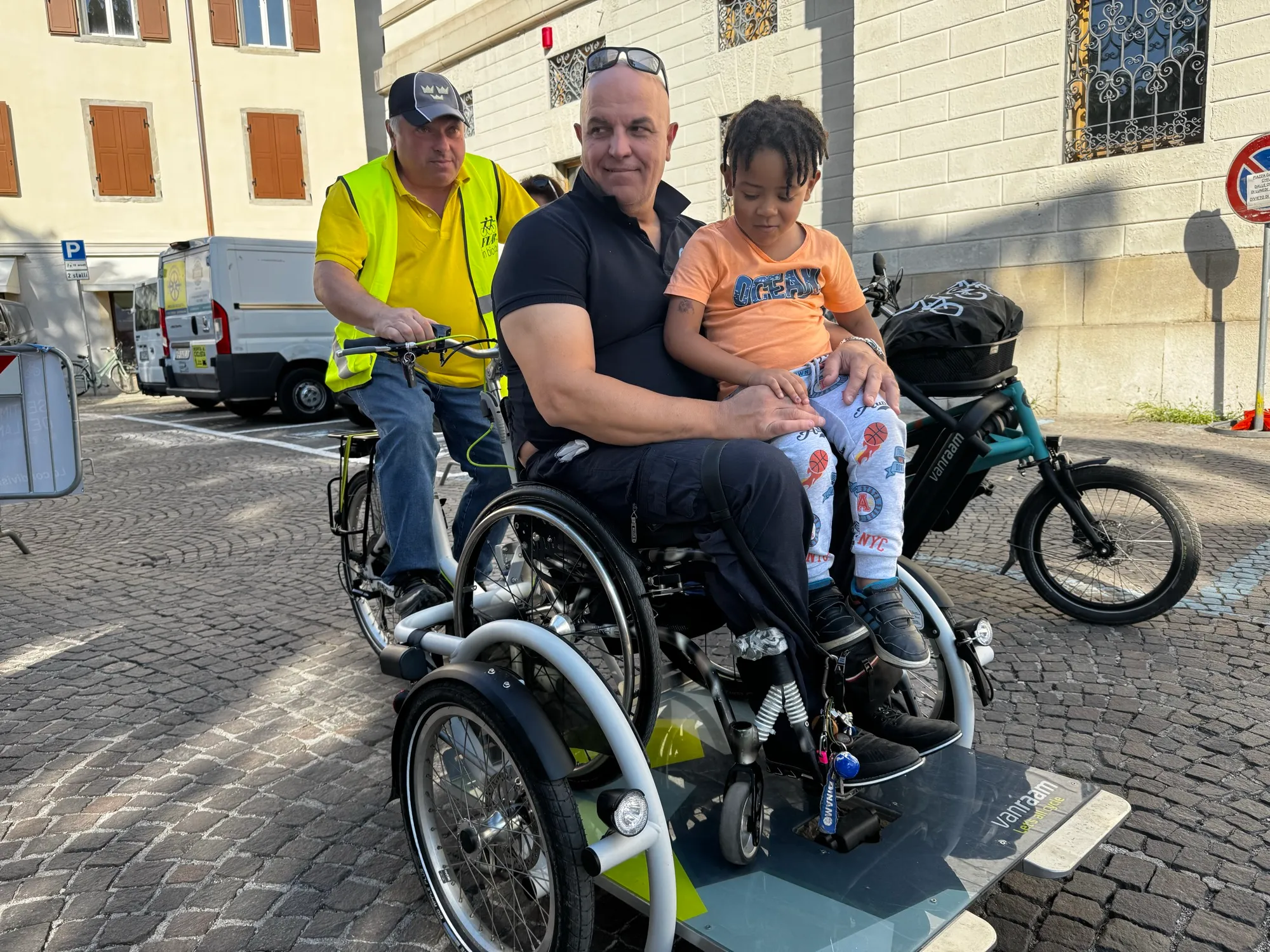 Un uomo in sedia a ruote agganciata a una bici Veloplus porta un bambino in braccio, in una piazza del centro storico.