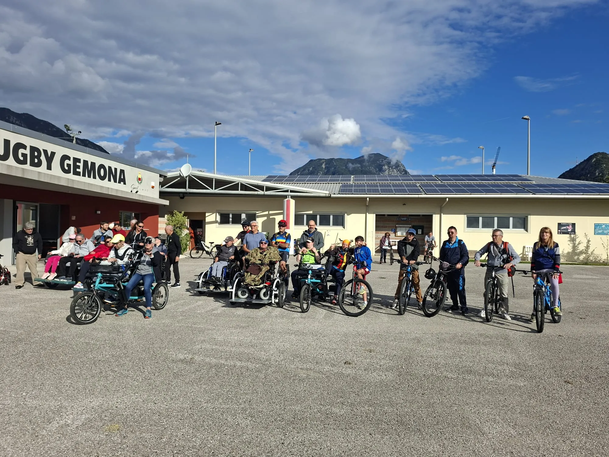 Un gruppo eterogeneo di persone, su biciclette e carrozzine adattate, si ritrova davanti a un circolo sportivo con le montagne sullo sfondo.