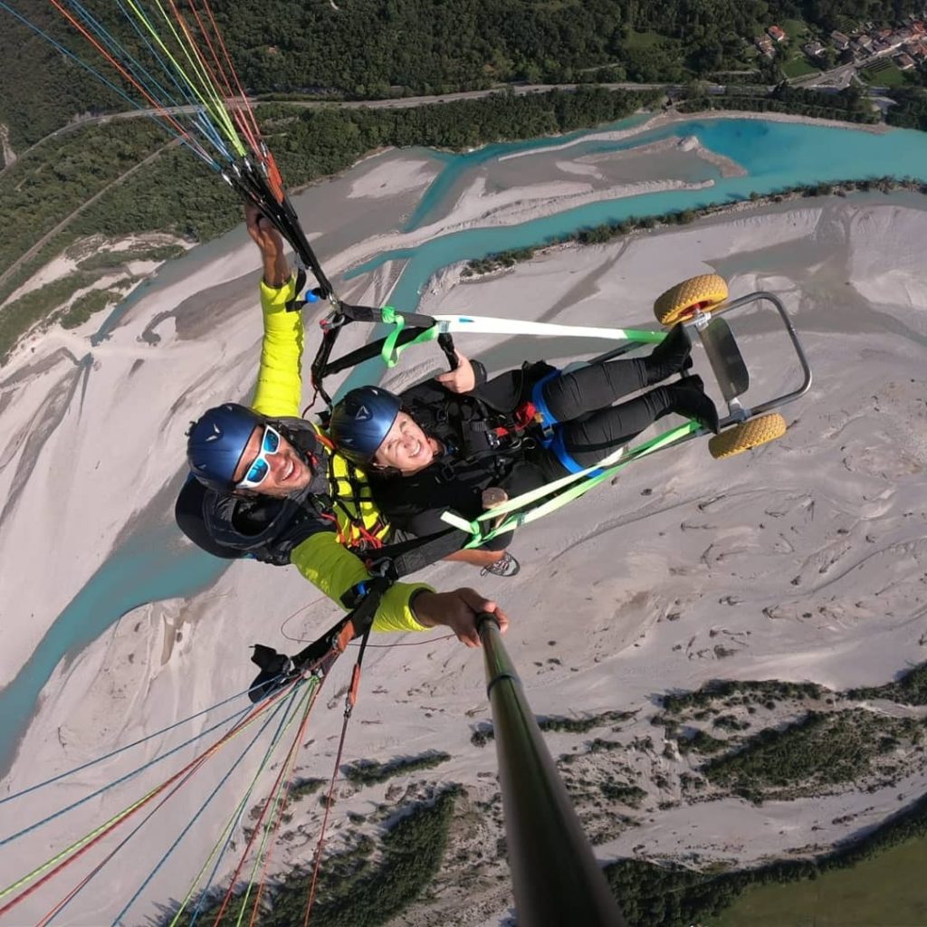 Due persone in volo con un parapendio biposto attrezzato con seggiolino a ruote, sopra un letto di fiume