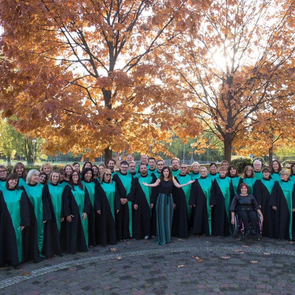 Un coro in abiti verdi e neri posa all'aperto tra alberi autunnali, con una corista seduta su una carrozzina in primo piano