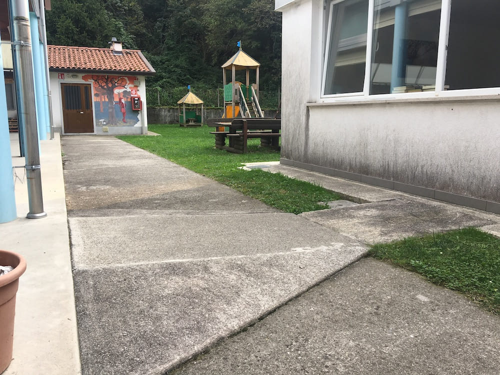 Percorso pavimentato tra un edificio con murale e la scuola, con un tratto di pavimentazione irregolare vicino al prato