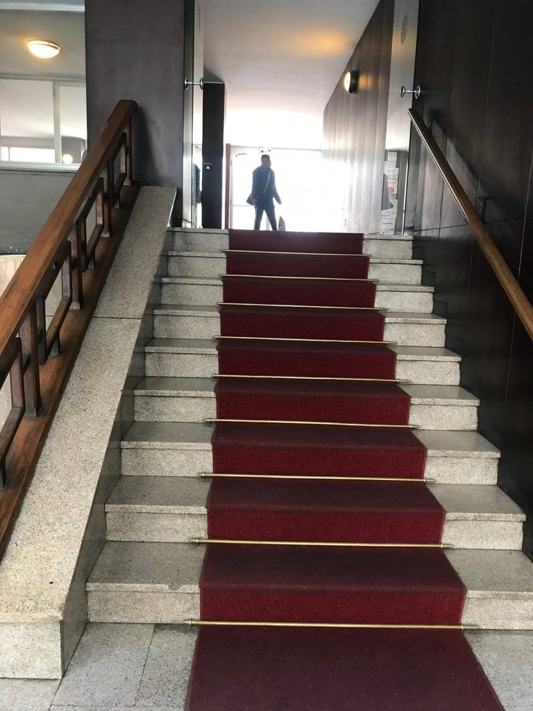 Scalinata interna con tappeto rosso e corrimano in legno su entrambi i lati, senza ascensore visibile
