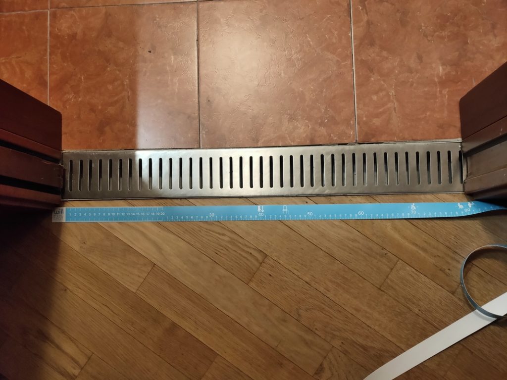 Misurazione con un metro a nastro della larghezza di una grata a pavimento tra una soglia piastrellata e un pavimento in legno