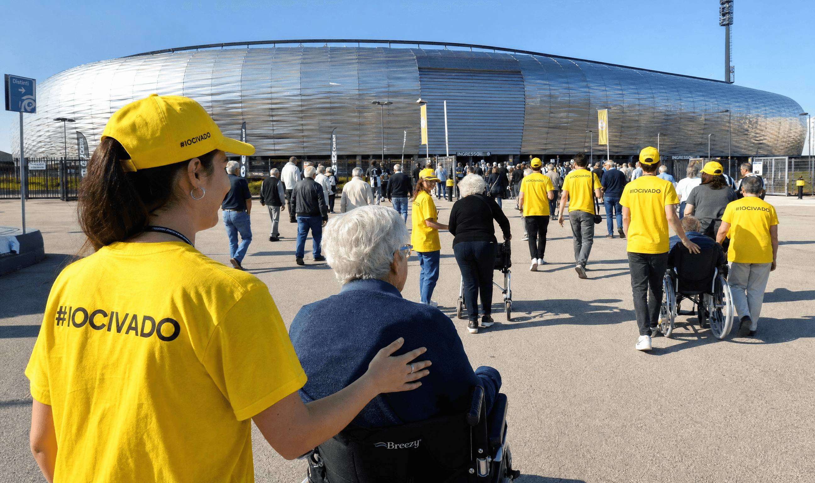 Una volontaria IO CI VADO con maglietta gialla "#IOCIVADO" accompagna una signora in
        sedia a rotelle verso l'ingresso della Dacia Arena, tra altri volontari e tifosi in fila
        per entrare.